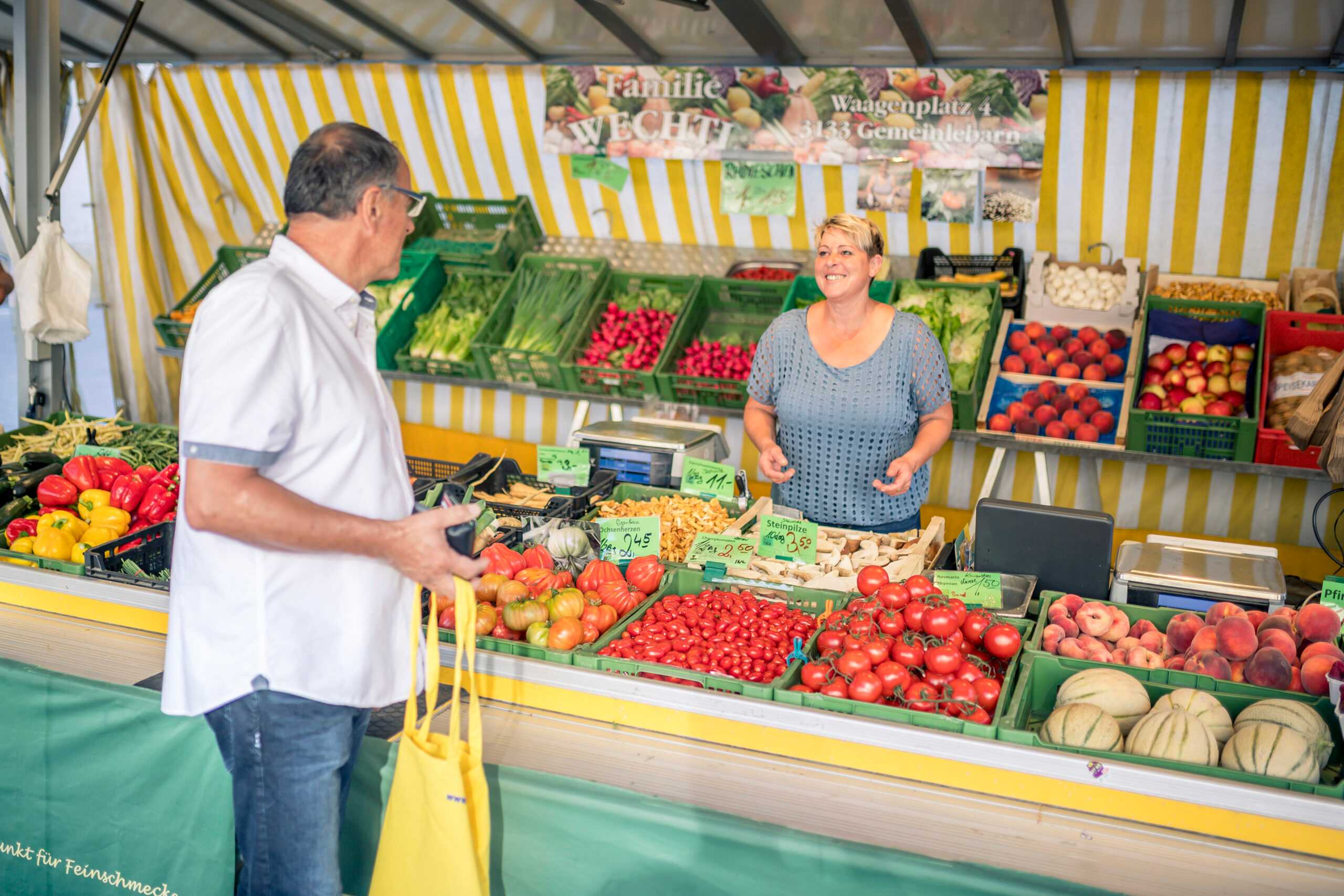 Das Foto zeigt einen Gemüsemarktstand mit buntem Gemüse, einer Verkäuferin und einem Käufer.