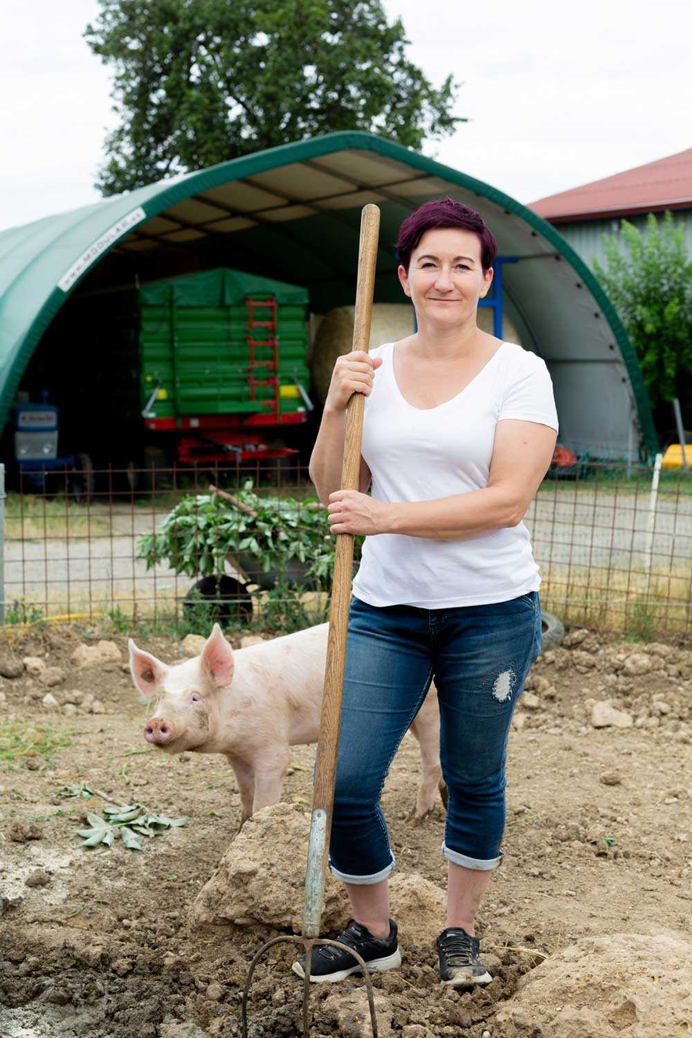 Das Foto zeigt eine selbstbewusste Bäuerin Jutta Schlager mit einer Mistgabel in der Hand, vor ihrem Bauernhof. Hinter ihren Beinen schaut ein junges Schwein hervor.